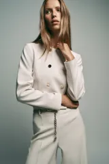 Blazer corta negra de cuello redondo y manga larga, con cierre frontal cruzado con botones dorados y falsos bolsillos delanteros.