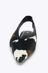 Zapato tipo bailarina destalonada en piel con pelo, con estampado animal print de vaca en blanco, marrón y negro. Tiene tira trasera con pieza elástica y acabado en punta. Suela plana de 1 cm de altura.