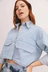 Camisa de denim azul con cuello clásico, cierre frontal con botones blancos, mangas largas con puños abotonados y dos bolsillos cargo con solapa en el pecho.