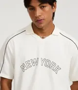 Remera tipo polo de hombre, color blanco crudo, con escote en V ribeteado en negro y ribetes negros en las costuras de los hombros. Presenta la inscripción arqueada "NEW YORK" en negro en el pecho.