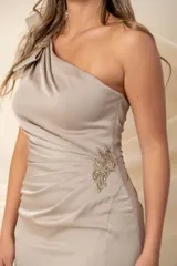 Vestido largo de fiesta color arena, de crepé, con escote asimétrico y un solo hombro. Presenta una moña con aplique bordado en el hombro y la cintura. Más largo atrás.