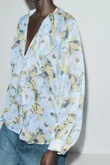 Camisa de cuello redondo y escote pico con estampado floral en tonos celeste, amarillo y negro. Manga larga abullonada acabada en puño. Detalle de volantes en el frente. Cierre frontal con botones.