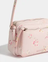 Bandolera pequeña de cuerina color rosa pálido con estampado floral multicolor. Tiene bolsillo frontal con cierre, correa regulable, cierre superior y tag metálico con logo.