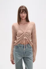 Sweater de poliéster color beige, con cuello en V, manga larga y frunces ajustables en el frente. Presenta un diseño de tejido calado.