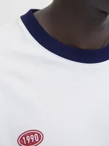 Remera blanca de manga corta y cuello redondo, con fit oversize y largo cropped. Tiene ribetes azul marino en el cuello y las mangas. Estampado frontal con la palabra "ORIGINALS" en negro y "studio" en cursiva, junto a detalles en rojo como "1990" y "COPENHAGEN CREATIVE DEPT.".