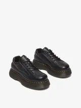 Zapatos Dr. Martens Buzz 5i de cuero negro con textura granulada, suela de plataforma gruesa con pespuntes amarillos, cierre de cordones y logo de la marca en relieve en el lateral.