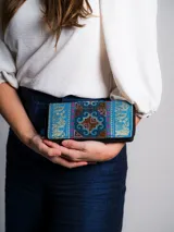 Billetera rectangular de tela bordada con motivos tribales en tonos celeste, azul, marrón y rosa. Tiene cierre con cremallera y tirador con pompón y cuentas de madera.