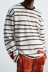 Camiseta relaxed fit de manga larga confeccionada en tejido compacto de algodón a rayas horizontales blancas y negras. Tiene cuello redondo y detalle de estampado de flores rojas en el bajo. Edición especial Zara x Otis Kwame Kye Quaicoe.