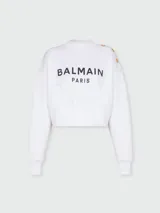 Sweatshirt blanco de corte cropped con cuello redondo y mangas largas. Estampado frontal con el logo de Balmain Paris en negro y botones dorados en el hombro.