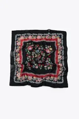 Pañuelo cuadrado negro con estampado floral en tonos rojos, rosas y verdes. Tiene un borde rojo con flores y un borde exterior negro con abalorios de colores.