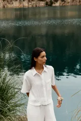 Camisa blanca de cuello solapa y manga corta, con bolsillo delantero y detalle de vainicas a tono. Cierre frontal con botones ocultos por solapa.