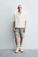 Camisa de denim de algodón, color beige claro, con corte relajado, cuello bowling y manga corta. Presenta cierre frontal con botones a presión y un pequeño bordado a tono en el pecho.