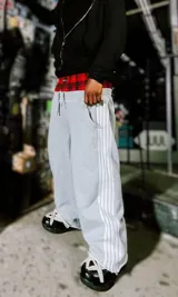 Pantalón deportivo gris jaspeado, estilo jogging, con corte ancho y tres franjas blancas verticales a lo largo de las piernas. Presenta un forro interior de tela escocesa roja y negra visible en la cintura.