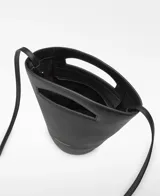 Bolso tipo capazo pequeño de piel ovina negra, con base cilíndrica y cuerpo que se ensancha hacia arriba. Presenta un asa corta troquelada en la parte superior y una correa larga ajustable y extraíble para llevar al hombro o cruzado. Tiene un compartimento principal con cierre de cremallera y el logo grabado en dorado en la parte inferior frontal.