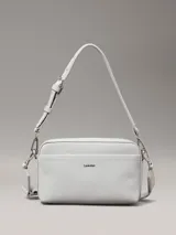 Cartera tipo camera bag de color blanco/gris claro, confeccionada en piel sintética con textura de monograma en el bolsillo frontal. Cuenta con dos correas desmontables: una ancha y otra fina, ambas ajustables. El bolsillo frontal tiene el logo de Calvin Klein en metal plateado.