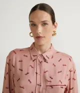 Camisa de viscosa color rosa con estampado de caballos en marrón. Tiene cuello camisero, cierre con botones, mangas largas, un bolsillo frontal y detalle de vivo contrastante.