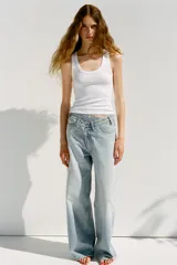Pantalón de jean celeste de tiro alto, corte wide leg y largo hasta el tobillo. Presenta un diseño asimétrico en la cintura con cierre frontal cruzado con cremallera y botones metálicos.