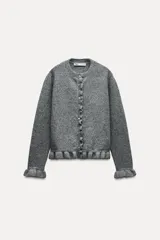 Cárdigan gris jaspeado de punto con cuello redondo y manga larga. Presenta volantes en el borde de los botones, en el bajo y en los puños.