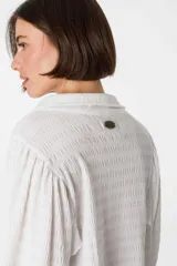 Camisa blanca de viscosa con cuello camisero, mangas largas abullonadas con puños elásticos y cierre frontal con botones. Presenta bordados negros con motivos florales en las mangas.