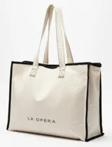 Bolso tipo tote bag de lona color crudo con ribete negro y asas largas. Estampado con el nombre de la marca en el frente.