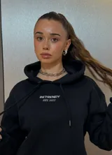 Hoodie negro de corte holgado con capucha y cordón ajustable. Estampado frontal con la leyenda "BE TRENDY EST. 2022" en color rosa.