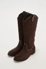 Botas vaqueras de caña media color marrón oscuro, confeccionadas en gamuza sintética. Presentan detalles de tachas plateadas alrededor de la abertura superior y en el borde del empeine, con un sutil bordado en la puntera. Tienen un taco bajo y plano.