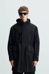 Parka negra de tejido técnico ligero, corte regular, cuello alto con capucha ajustable, manga larga con puños ajustables, cintura ajustable con cordón interno, bolsillos de solapa en la cadera, abertura en la espalda y cierre frontal con cremallera oculta y botones a presión.