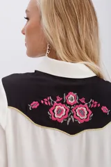 Camisa blanca de viscosa con canesú negro bordado con motivos florales en rosa y dorado. Tiene mangas largas con puños con snap metálico, acceso frontal con snaps metálicos y vivos en lurex dorado en canesú y en bolsillos ojales decorativos.
