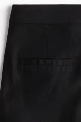 Pantalón sastrero tobillero negro de tiro alto con cierre y pretina prolongada con botón. Presenta bolsillos al bies, bolsillos traseros decorativos y piernas amplias con pliegues adelante y atrás.