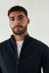 Campera tejida azul marino con cuello alto, cierre frontal y puños acanalados con detalle de línea roja.