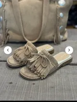 Sandalias planas tipo mule, color beige, con tira ancha adornada con flecos y lazo de gamuza.