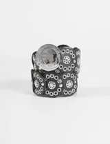 Cinturón ancho de cuero sintético negro, compuesto por secciones circulares unidas, decorado con múltiples ojales metálicos plateados y un dije central grande con relieve de un jinete a caballo. El cierre es mediante un broche metálico grande con el mismo diseño de relieve.