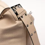 Bolso tipo hobo color beige con textura granulada y correa de hombro ajustable. Cierre superior con cremallera y tiras decorativas laterales con detalle de nudo.