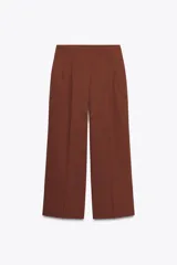 Pantalón marrón de tiro alto con bolsillos delanteros y falsos bolsillos traseros con tapeta. Presenta pliegues en la parte delantera y cierre frontal con cremallera y gancho metálico.