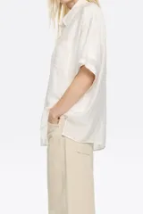 Camisa oversize blanca con rayas verticales finas en gris oscuro, cuello camisero, manga corta ancha, cierre frontal con botones y bolsillo de parche en el pecho. Confeccionada en viscosa y poliamida.