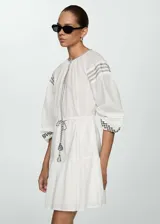 Vestido corto blanco de algodón con bordados y detalles calados. Tiene cuello redondo, mangas largas con puños elásticos, cintura elástica con lazo y diseño evasé.