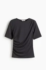 Remera negra de punto crepé con escote redondo y mangas cortas. Diseño ajustado con frunces en un hombro y en el lado opuesto de la cintura para crear un leve efecto drapeado.