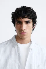 Camisa celeste a rayas verticales, de corte relaxed fit, confeccionada en lino y algodón. Tiene cuello abotonado, manga larga con puños abotonados, bolsillo de parche en el pecho y cierre frontal con botones.