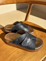 Sandalias de cuero color azul marino con plantilla dorada y suela marrón. Presentan una tira ancha cruzada con doble remache dorado.
