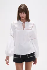 Blusa holgada de algodón color rosa pálido, con escote redondo y lazo ajustable en el centro. Presenta detalles de encaje calado alrededor del escote y en los puños de las mangas largas abullonadas.