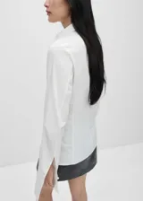 Camisa blanca de algodón con cuello camisero, corte recto, mangas largas con pliegues y puños abotonados.