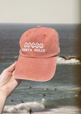 Gorro tipo dad hat color terracota desgastado, con visera curva. Presenta bordado frontal en hilo blanco con un patrón ondulado y el texto "COSTA DULCE" debajo.