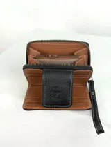 Billetera pequeña tipo clutch con correa de muñeca, confeccionada en cuero negro texturizado y un panel central de pelo (pellejo) en tonos blanco y negro. Presenta costuras blancas de contraste y un cierre de solapa con broche.