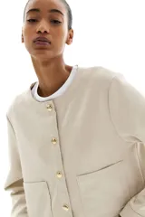 Saco corto color beige de tejido de punto con acabado cepillado suave, cuello redondo, cierre frontal con botones metálicos dorados y bolsillos de parche delanteros.