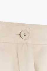 Pantalón color beige de tiro alto, corte ancho y largo midi, con pinzas delanteras, bolsillos laterales y traseros. Cierre frontal con cremallera y botón.