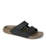 Sandalias tipo slide de dos tiras ajustables con velcro, marca Reef. La capellada es negra y la suela es de goma EVA en color marrón claro. El nombre de la marca está grabado en relieve en la tira lateral.
