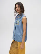 Camisa sin mangas de denim liviano color camel, con cuello camisero y botones frontales. Presenta bordados temáticos marinos en el pecho, incluyendo un sol, olas, una gaviota y vida marina.