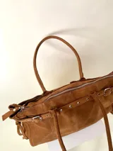 Bolso grande de cuero marrón con textura tipo cocodrilo, estructura rígida y asas para llevar al hombro. Tiene una tira con tachas y hebillas en cada extremo, también lleva lazos en los costados.