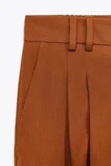 Pantalón color marrón confeccionado en hilatura con mezcla de lyocell y algodón. Tiro medio, cintura con trabillas, bolsillos laterales, pinzas en delantero y raya planchada. Cierre frontal con cremallera, botón y ganchos metálicos.