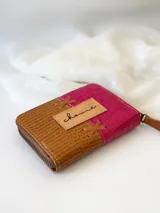Billetera pequeña de cuero color fucsia y marrón con textura tipo piel de cocodrilo. Tiene cierre metálico y detalle de tiras de cuero cruzadas en el frente.
